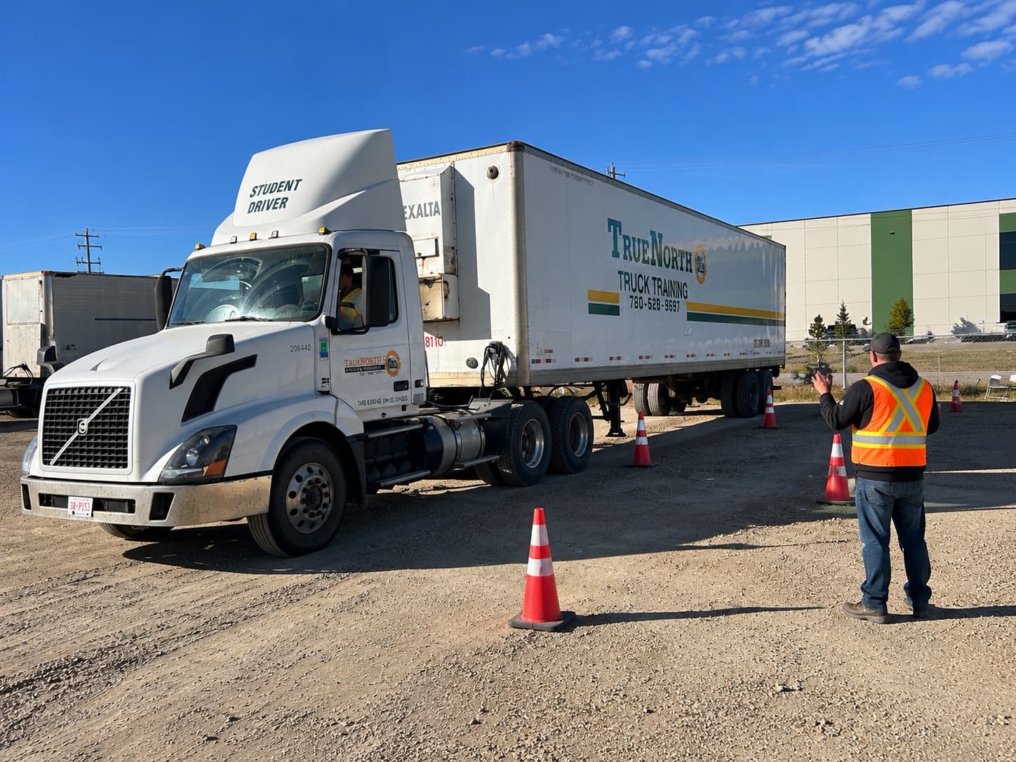 Instructor in a safety vest guiding a student driver maneuvering a tractor-trailer through a cone course